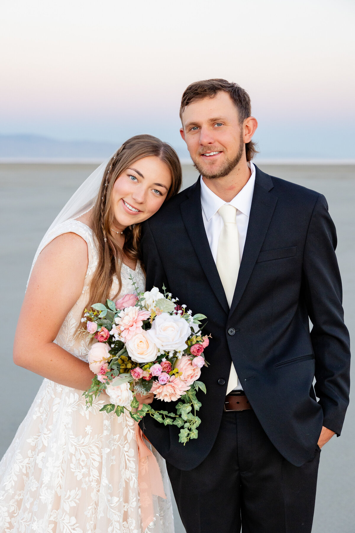 Bride leans her head on the groom’s shoulder, holding a bouquet, both smiling with a soft sky behind them.