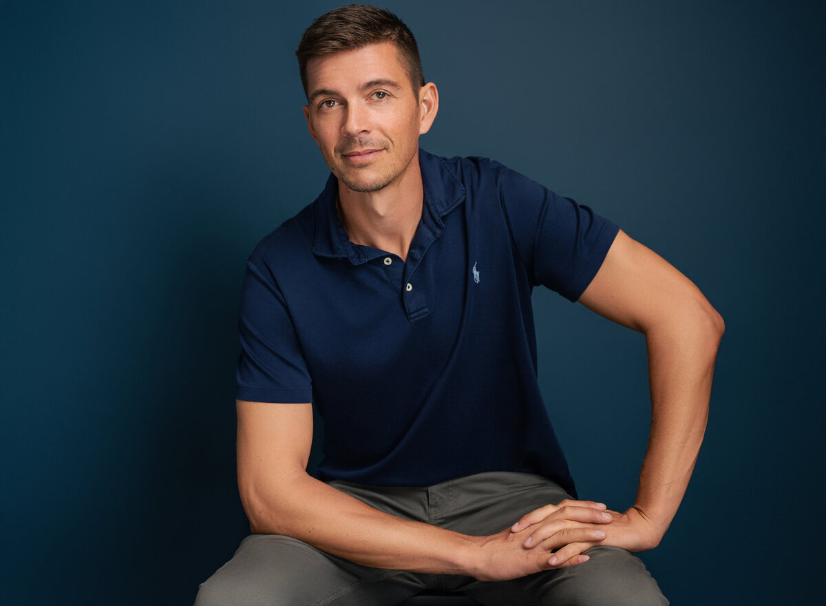 Man in a navy polo shirt, sitting and posing with one arm resting on his knee