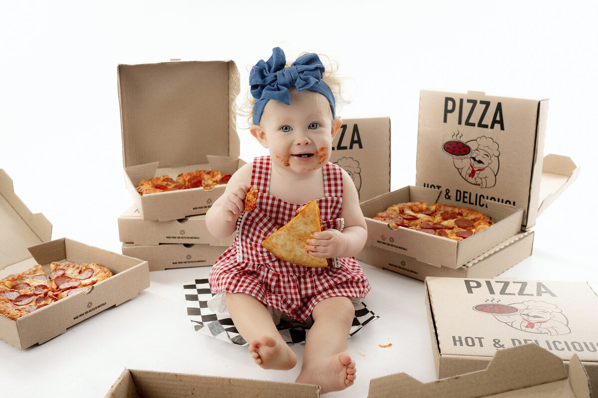Toddler in a checkered outfit with a bow, surrounded by pizza boxes, holding a pizza slice.