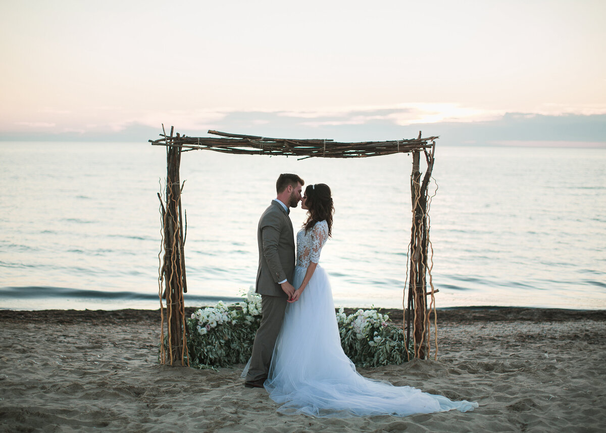 The bride and groom stand under a rustic archway on a sandy beach with the sunset beyond.