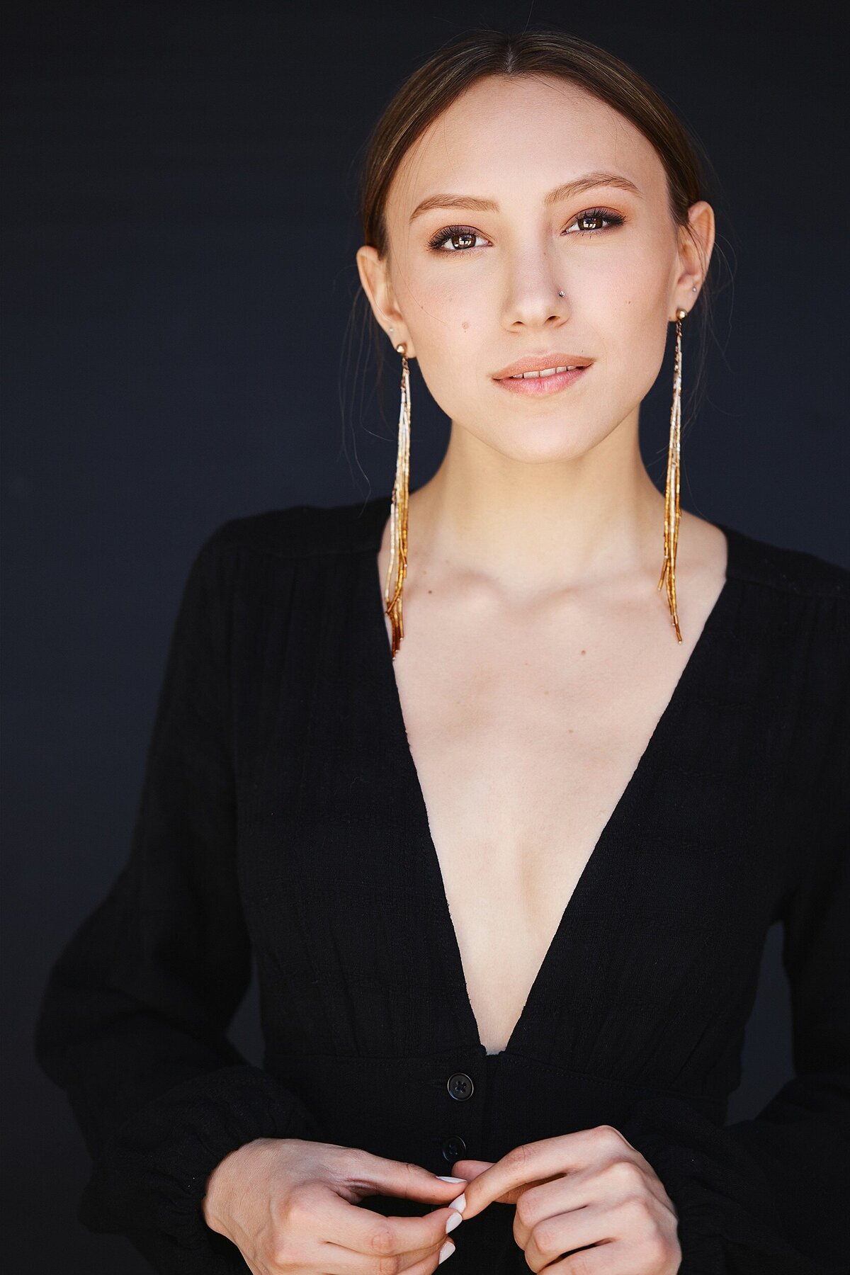 Studio portrait of a young woman with long earrings and natural makeup, wearing a black V-neck top, captured against a dark backdrop.