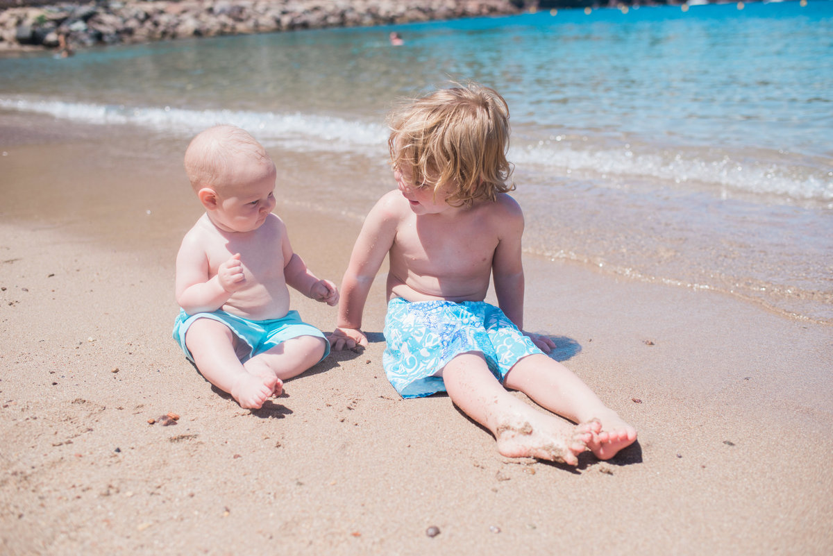 little boy brother sitting on beach with blue water