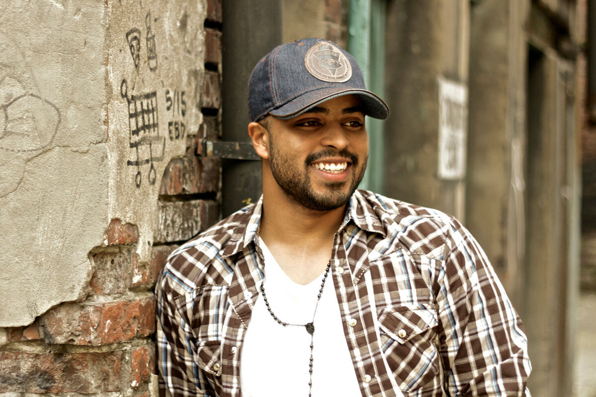 Country Music Photo Jojo Mason standing leaning against brick wall wearing plaid shirt and baseball cap smiling