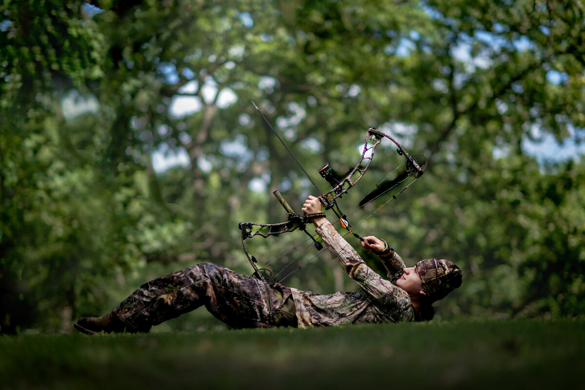 A hunter lays in the grass aiming his bow to a tree in the forest