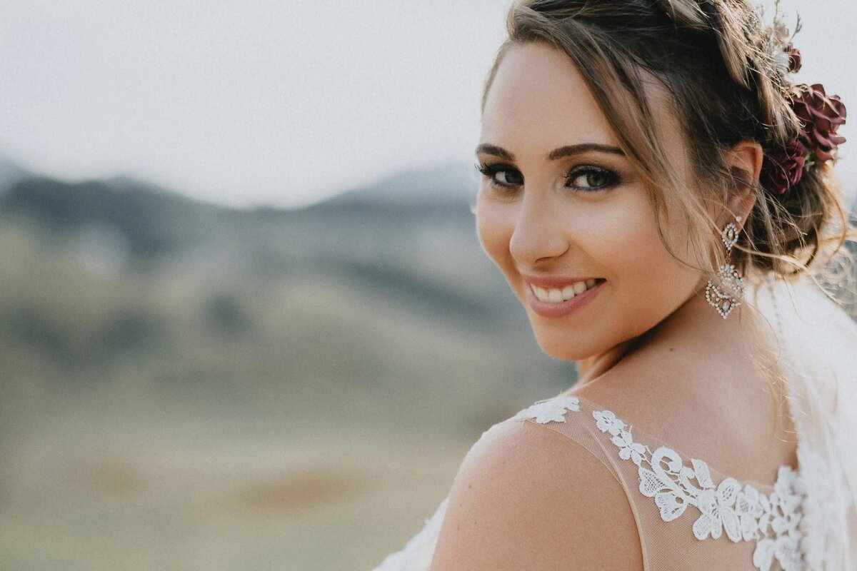 A portrait of a smiling bride with her hair styled up and a delicate lace dress, with a serene landscape in the background.