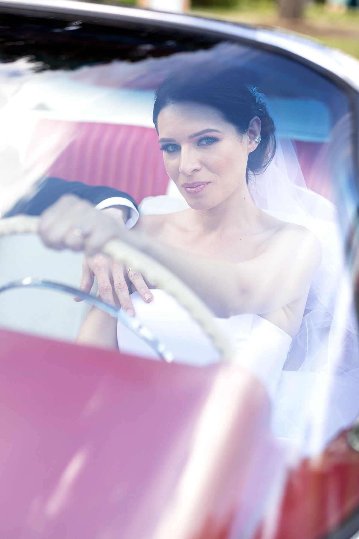 Bride in white wedding dress  posing in the driver seat of vintage old red convertible car with groom in passenger seat