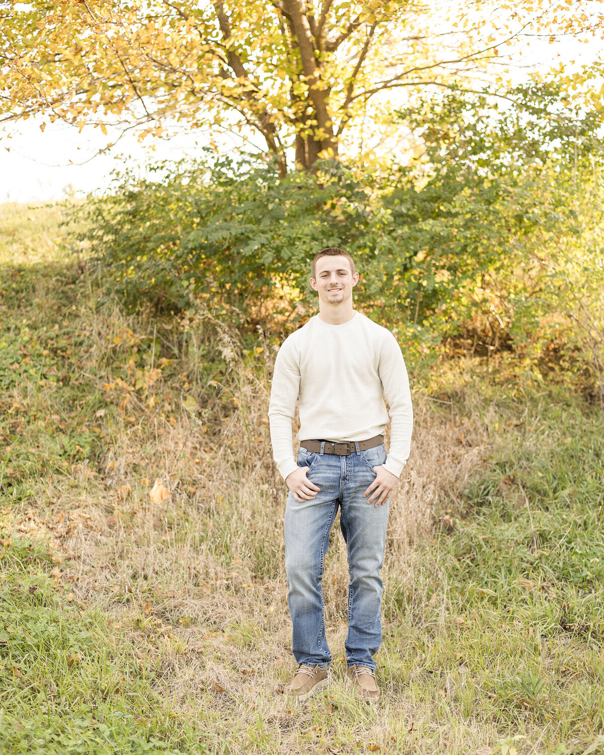high school senior male standing in grass, with a tree behind him, wearing blue jeans, light brown dress shoes, long sleeve light tan shirt, a brown belt, thumbs in his pockets smiling at the camera