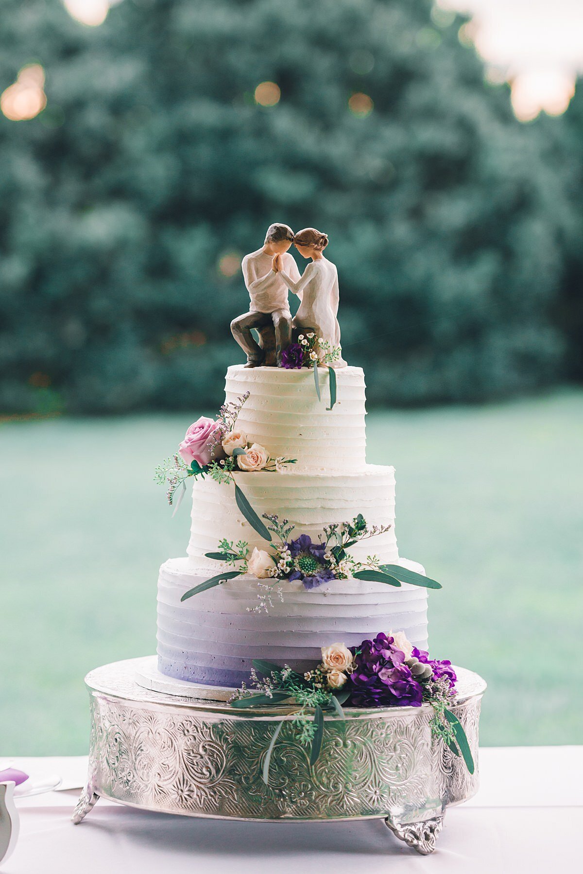 Ombre three tiered wedding cake with pink and purple flowers sitting on a silver cake stand topped with a bride and groom topper
