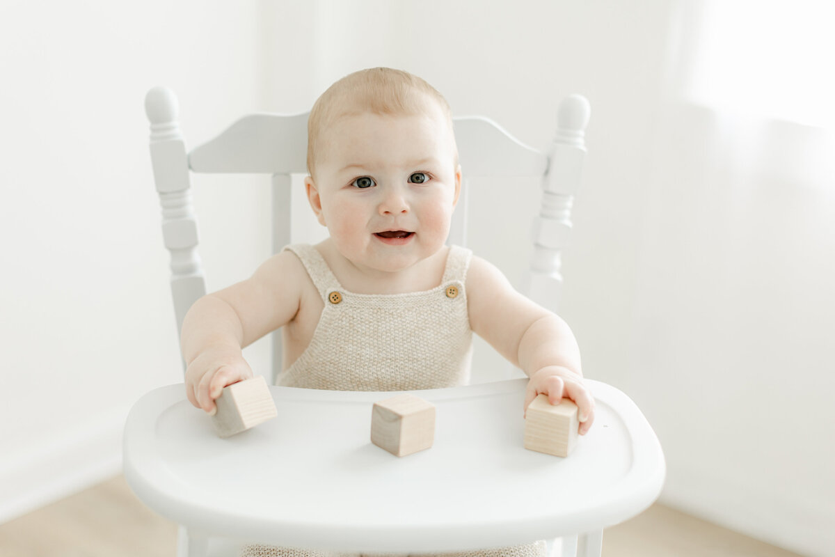 1 year old baby boy playing with wooden blocks sitting in a white wooden  highchair photographed by Philadelphia Portrait Photographer Tara Federico