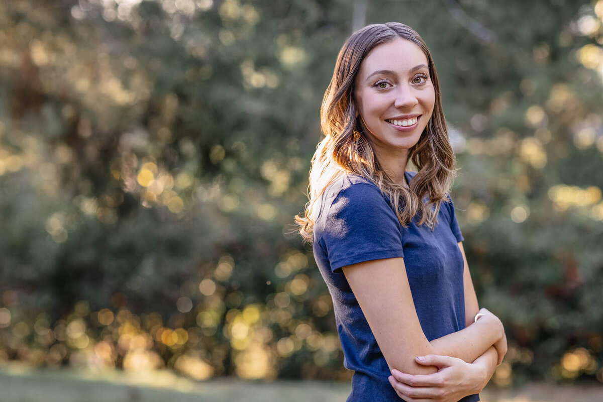 A beautiful young woman wearing a navy blue shirt standing in a green park, holding her arms and smiling sideways at the camera.