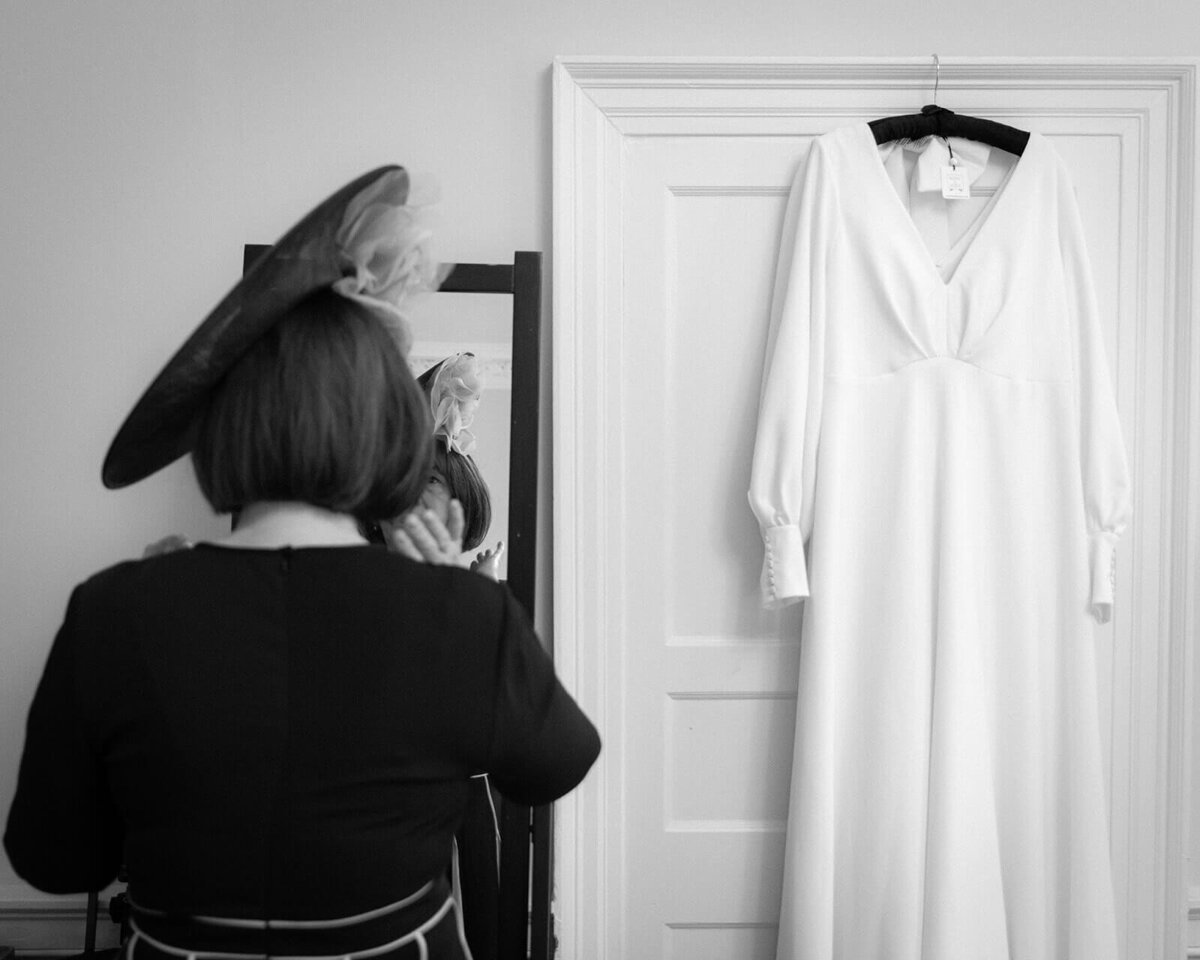 A mother of the bride puts the final details together before a wedding. She adjusts her hat in the mirror with a wedding dress hanging to her right.