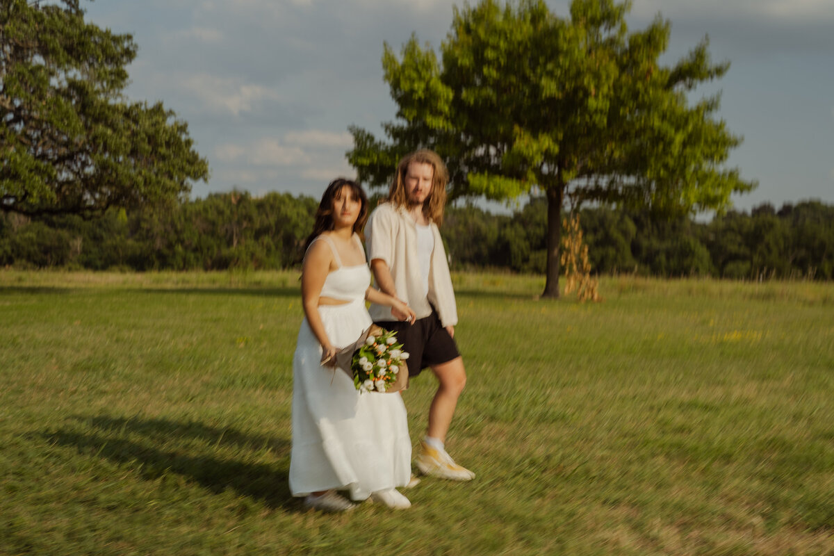 A candid and warm springtime photo of a couple walking hand in hand through the park near White Rock Lake in Dallas. The couple shares a peaceful moment with a bouquet in hand, surrounded by the lush greenery of the park, perfect for engagement photography sessions in Dallas and Fort Worth, Texas.