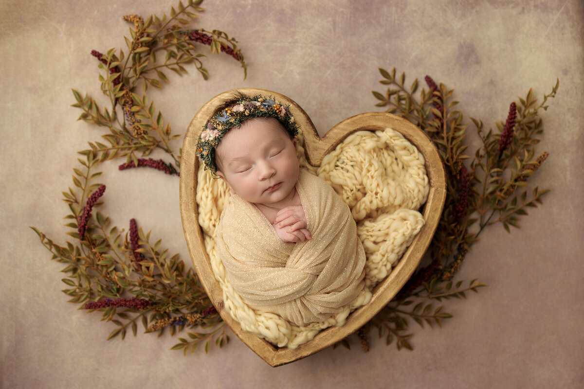 Newborn baby swaddled in a beige blanket, sleeping peacefully in a heart-shaped wooden bowl, surrounded by dried flowers and greenery, wearing a floral headband.