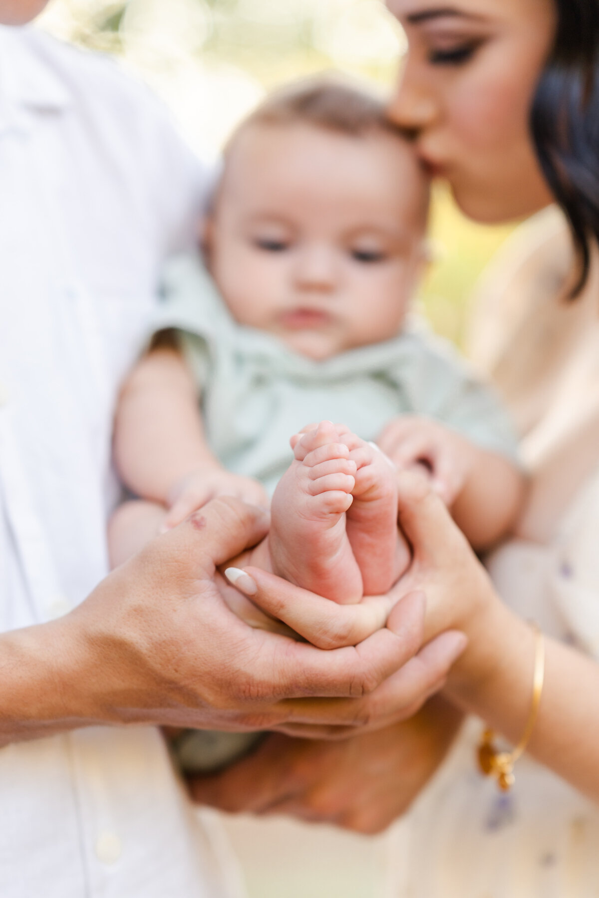 A baby in a soft green jumper rests between his mom and dad as they hold his cute feet and the mother gently kisses his head photographed by Bay Area Family Photographer, Light Livin Photography.