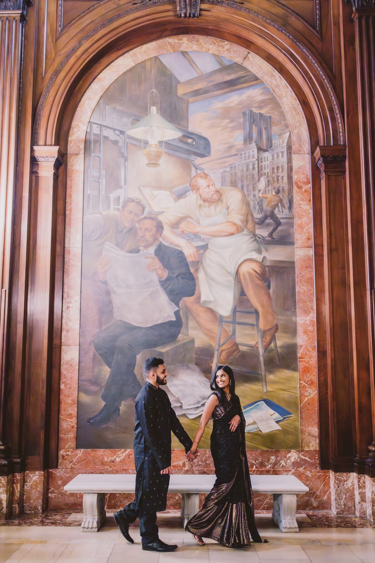An engaged couple walk near a mural indoors at the new york public library inside in indian clothing