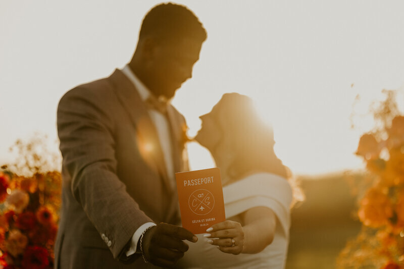 Mariés en extérieur dans un rayon de soleil couchant, fleurs rouges en arrière plan, tenant au premier plan un passeport rouge pour la séance photo mariage.