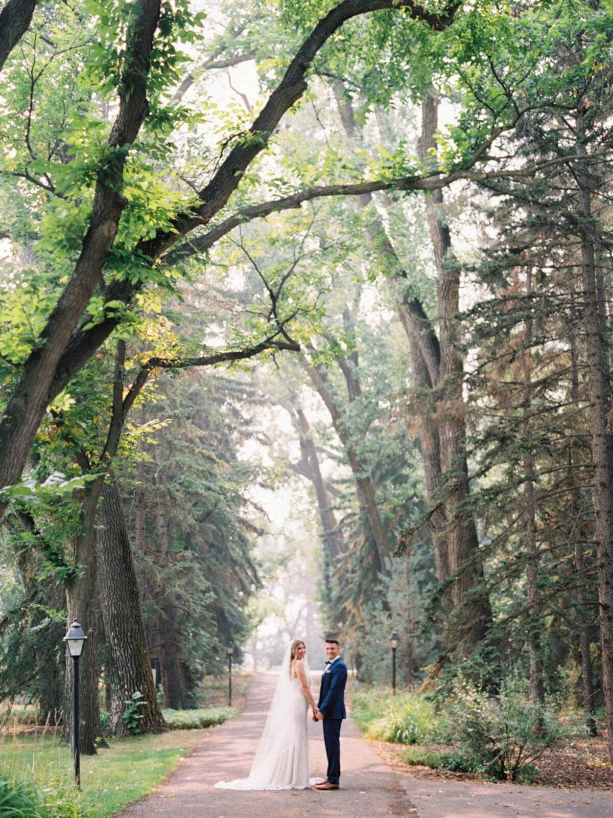 Bride and groom on the tree-lined path at The Norland Historic Estate, a classic vintage wedding venue in Lethbridge, AB, featured on the Brontë Bride Vendor Guide.