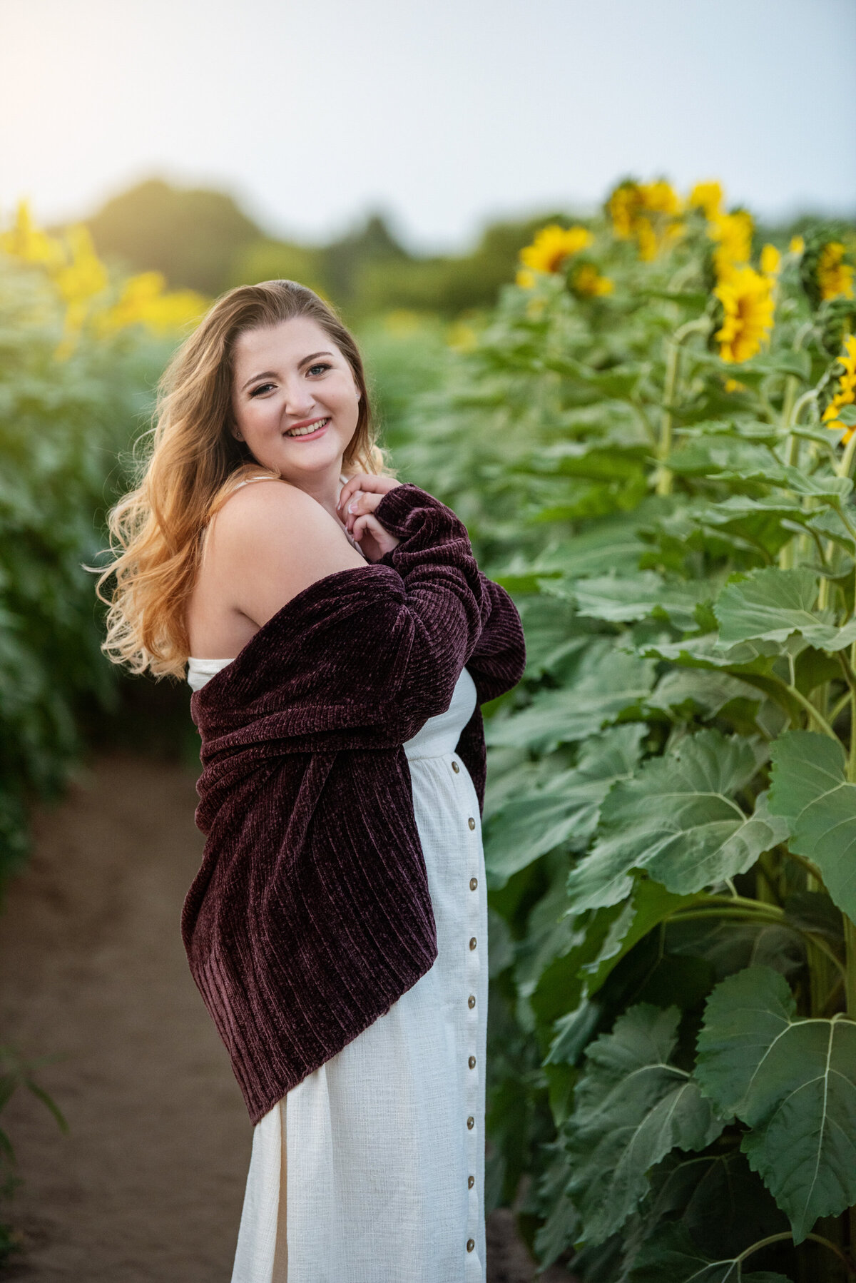 girl wearing white dress and maroon cardigan in row of sunflowers