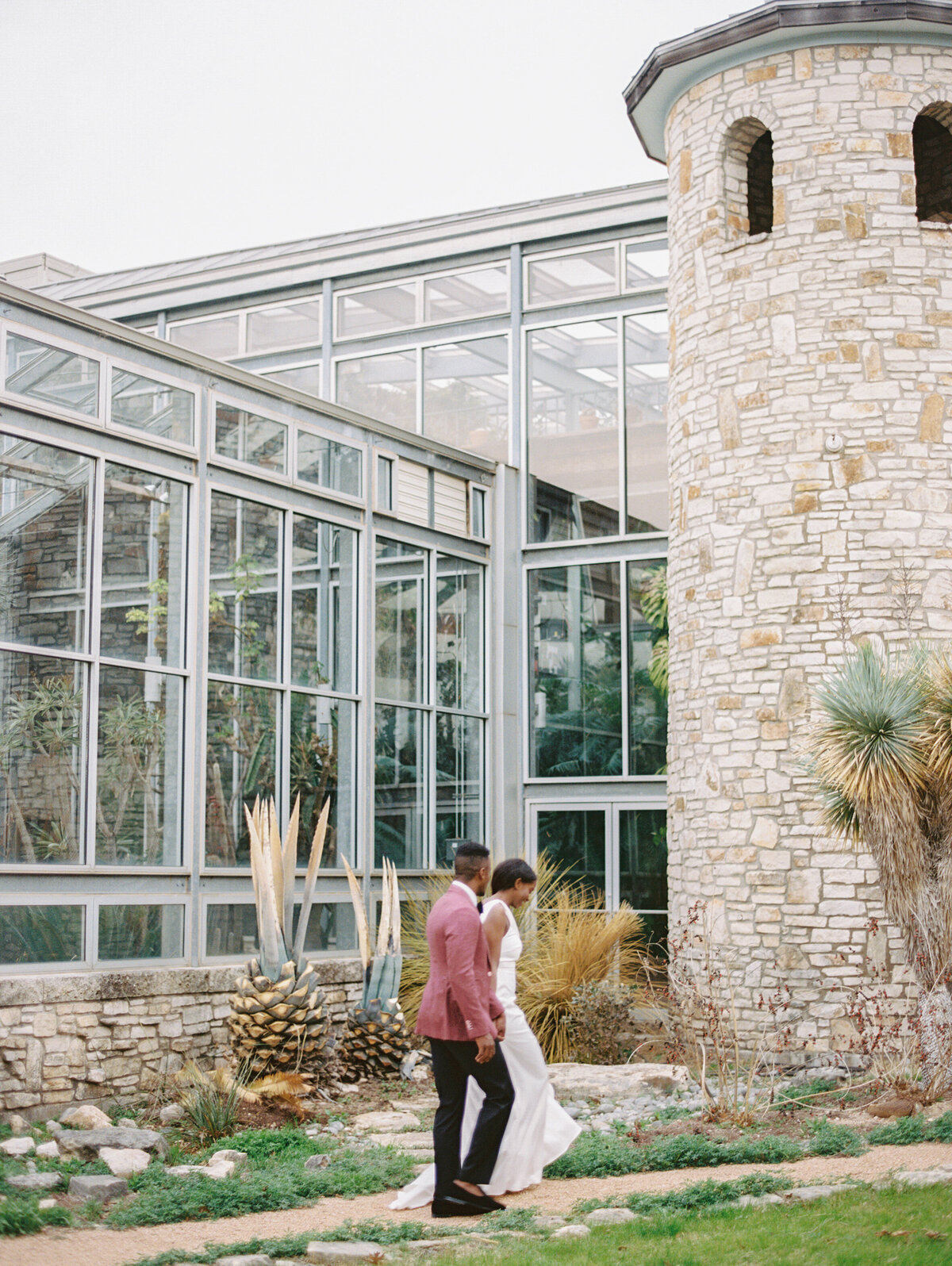 groom in a pink jacket leads bride past the greenhouse at driftwood