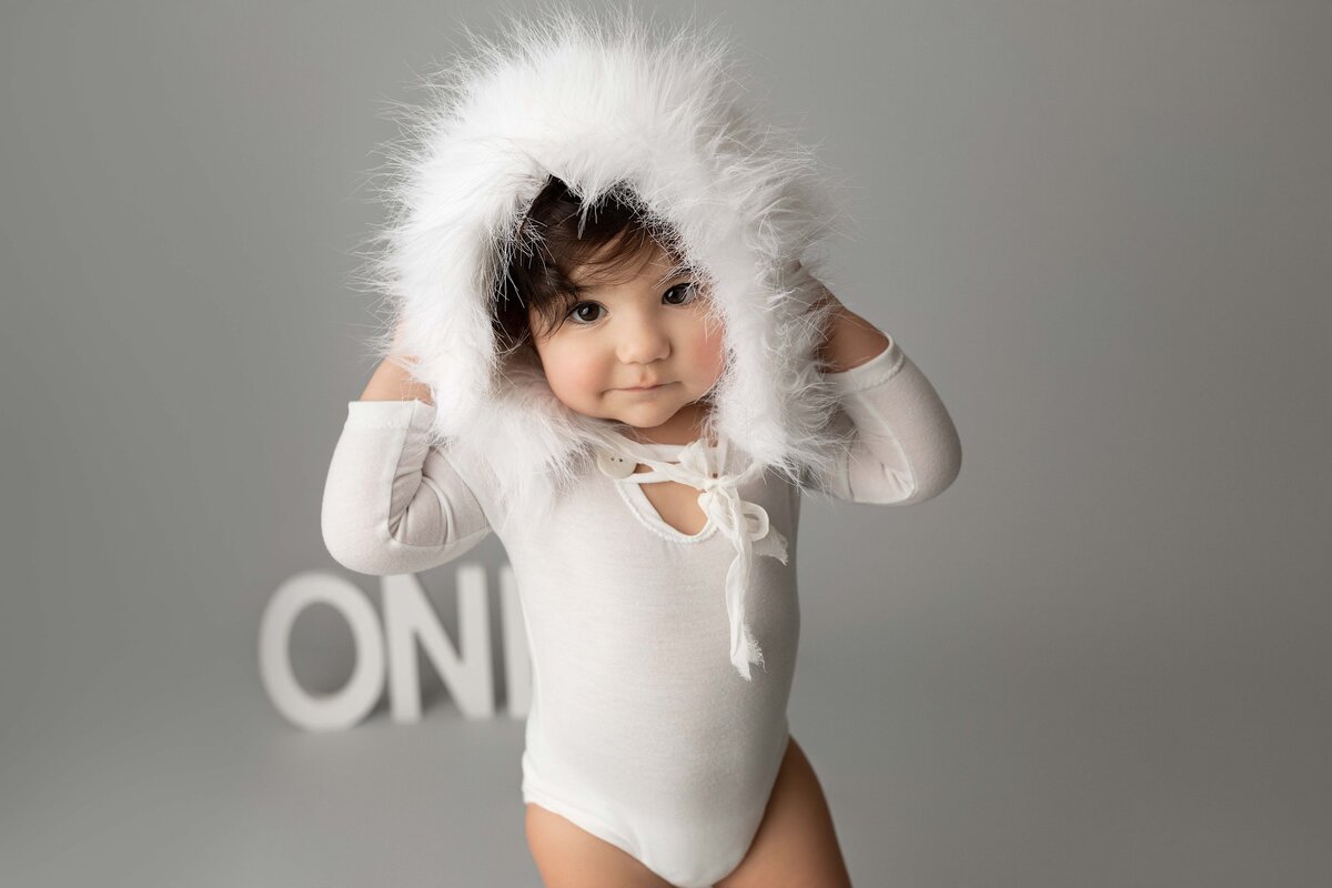First birthday portraits in London, Ontario studio - baby in white onesie and faux fur trimmed hat looking at the camera. Both of baby's arms are touching the fur. The letters o-n-e appear in the background.