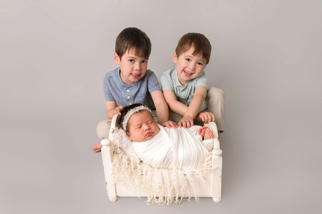 Two young brothers posing behind their newborn baby sister in white wooden bed prop during studio photography session