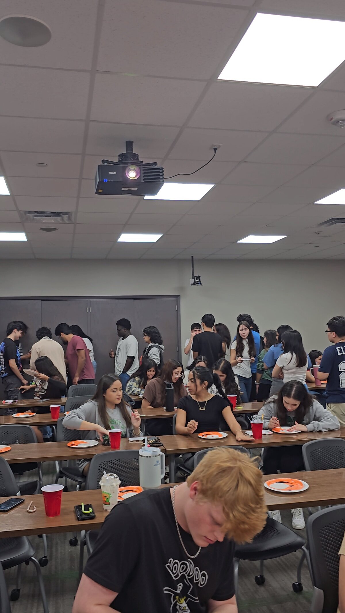 The vibrant world of creativity at Soleil Vida Studio's giant art room. A group of young individuals eagerly lines up to gather materials for their graphic pieces. Some are already seated at large wooden tables, fully immersed in the artistic process.