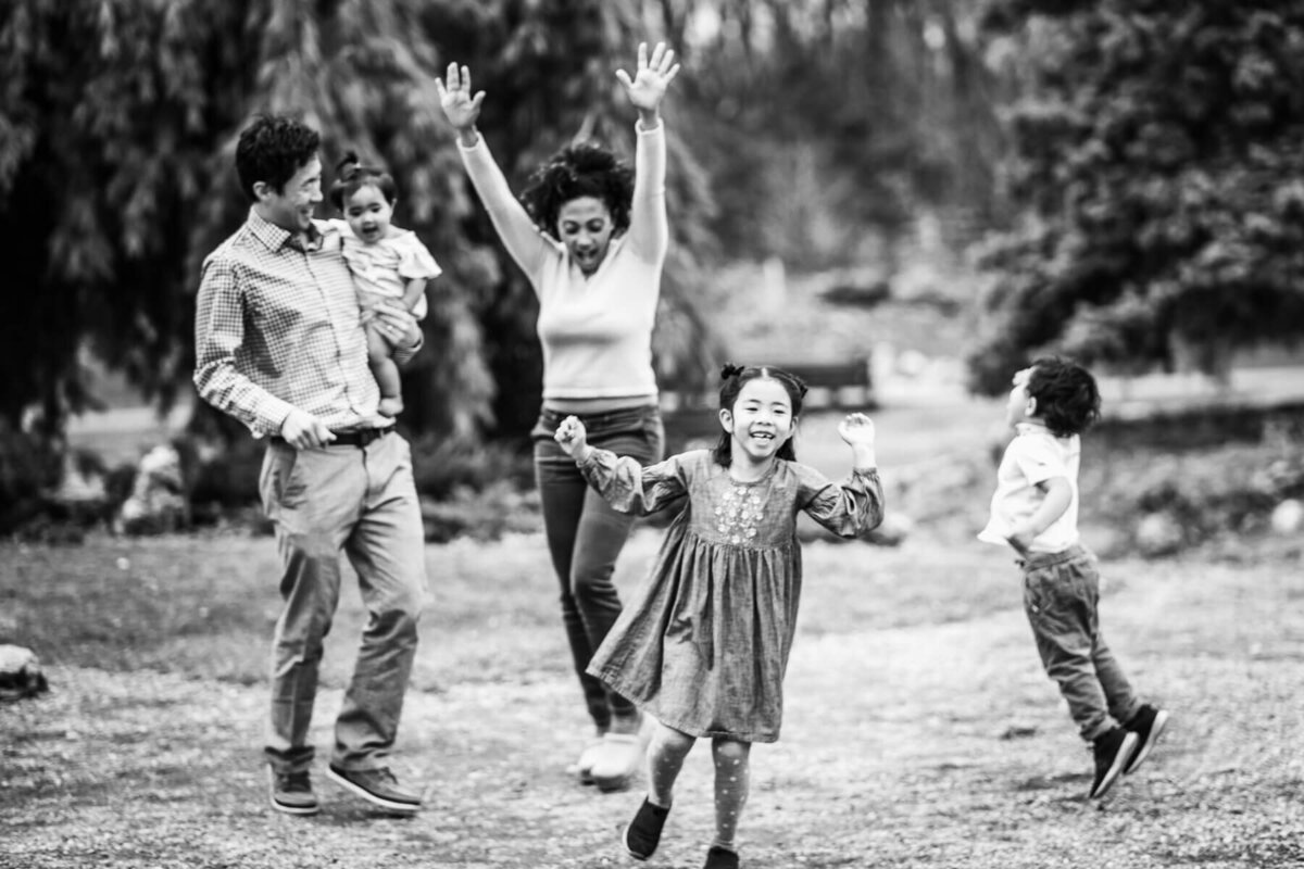 A black and white image of a young family dancing and jumping during a family photoshoot at the Lyndale Park Rose Gardens in Minneapolis, Minnnesota.