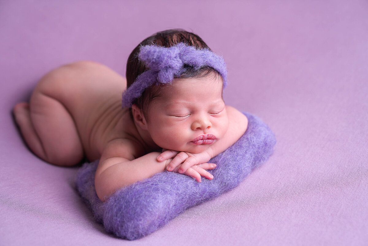 Bare baby girl is sleeping  on her belly with her hands folded underneath her chin for newborn photoshoot. She is wearing a purple headband and her arms are laying atop of a purple pillow. Captured by Main Line photographer Liza Nicole.