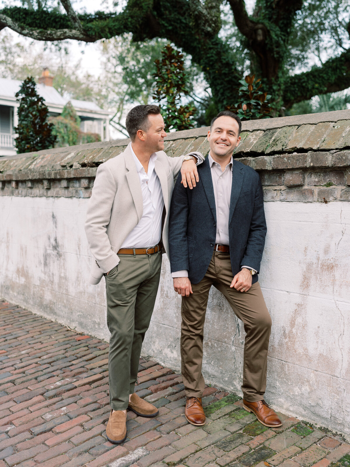 A couple enjoying a moment of togetherness leans casually against a historic wall. Their stylish attire and relaxed expressions enhance the engaging atmosphere of the scene in Charleston, SC.