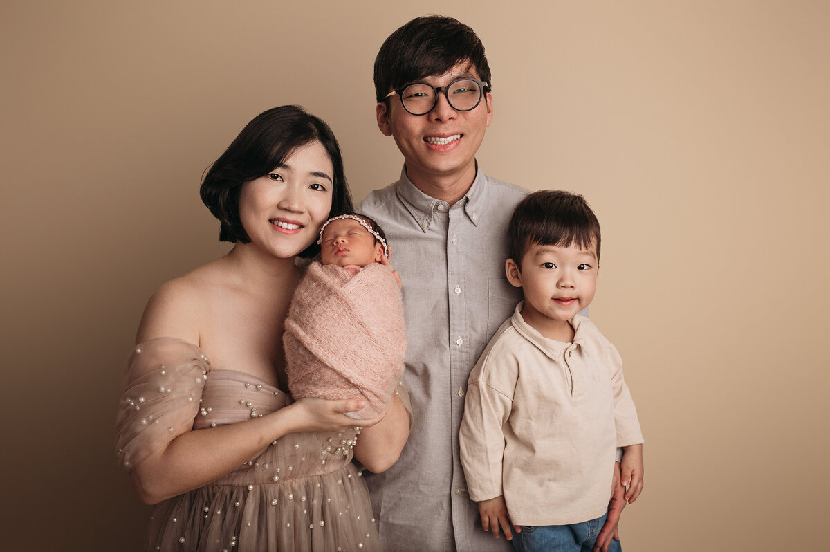 beautiful family of four from China  together with mom smiling wearing a cream pearl dress while holding newborn baby girl swaddled in  a pink knit wrap standing next to smiling dad wearing glasses and grey button down shirt standing next to toddler big brother smiling at the camera in front of cream backdrop
