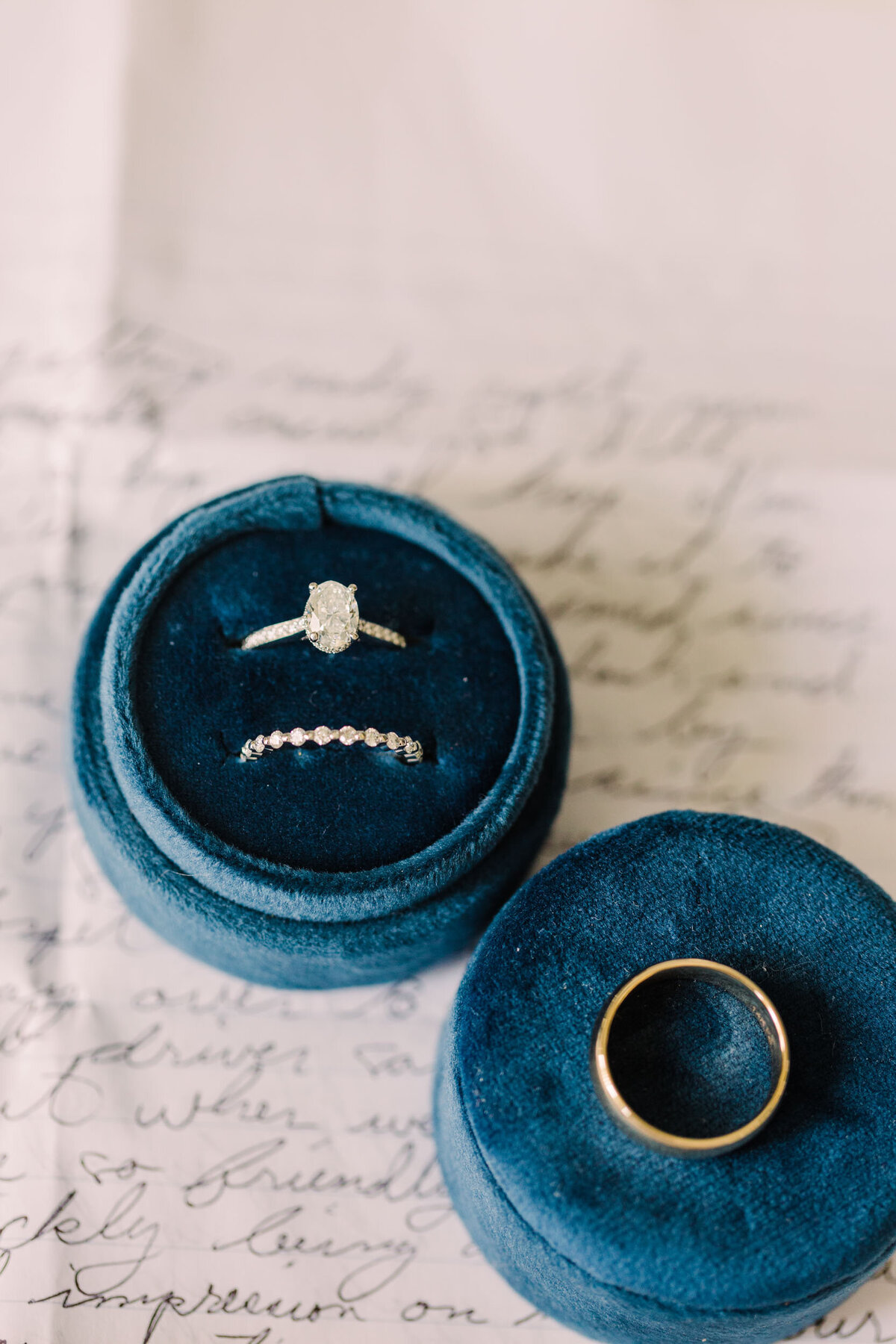 A close-up wedding ring detail with gold bands placed inside blue velvet boxes, resting atop a handwritten letter from the groom.