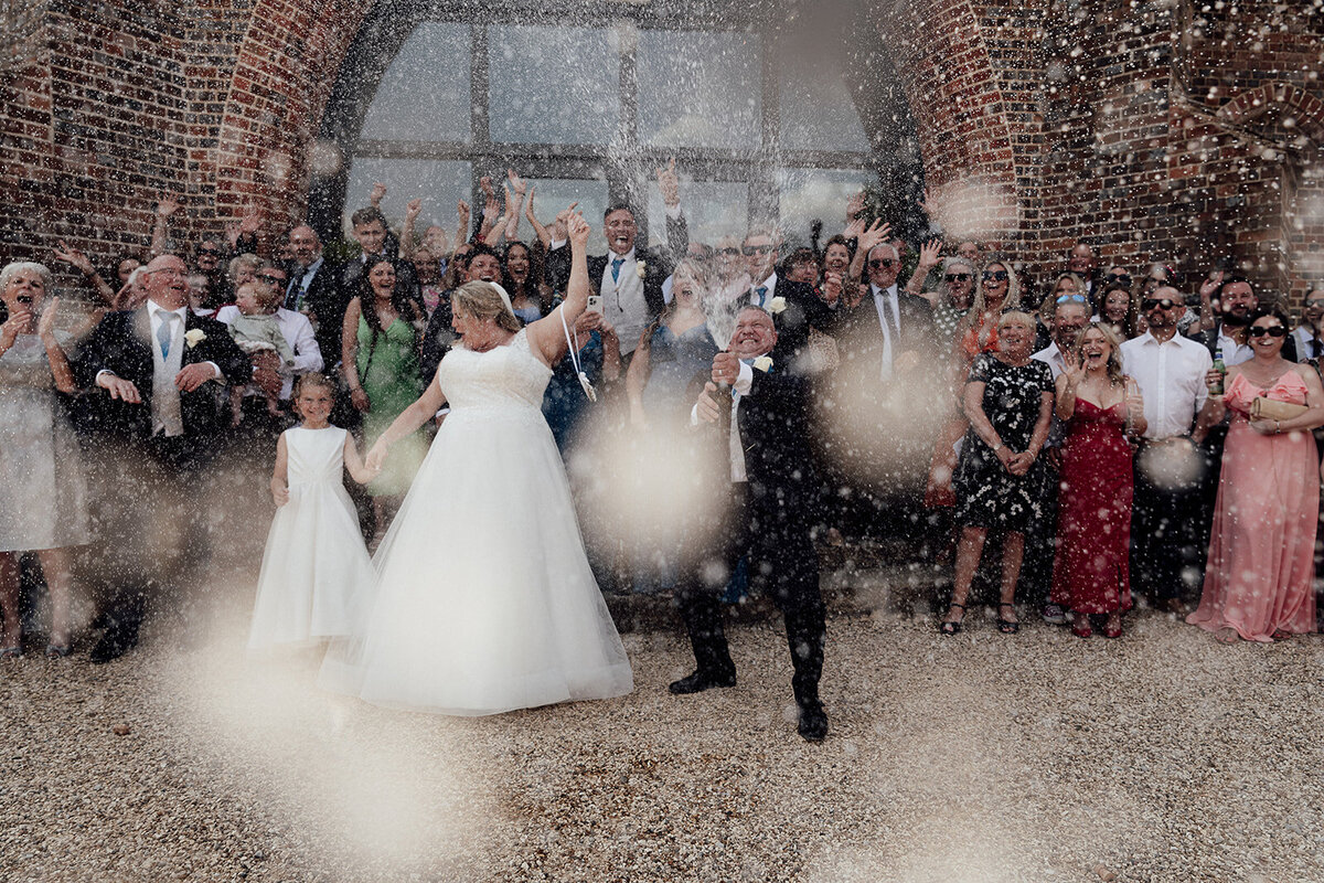 A wedding party spray champagne towards the camera as they cheer in a tight group.