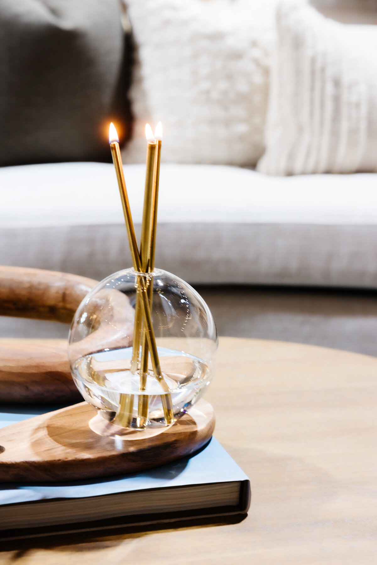 Close-up of a decorative candle arrangement with two lit candles in a glass holder, placed on a wooden tray in a modern, minimalistic living room setting.