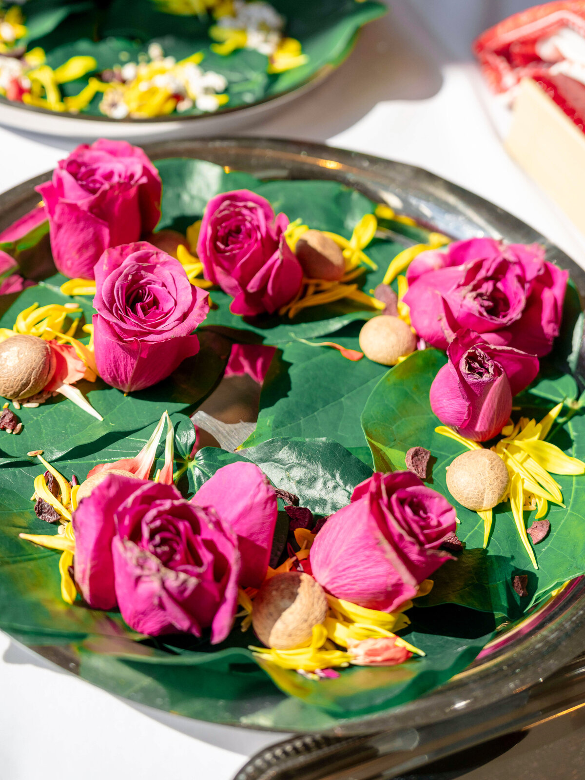 A decorative arrangement on a silver platter featuring vibrant pink roses, yellow and white flowers, and small round beige objects nestled on a bed of green leaves. The background shows part of another similar arrangement.