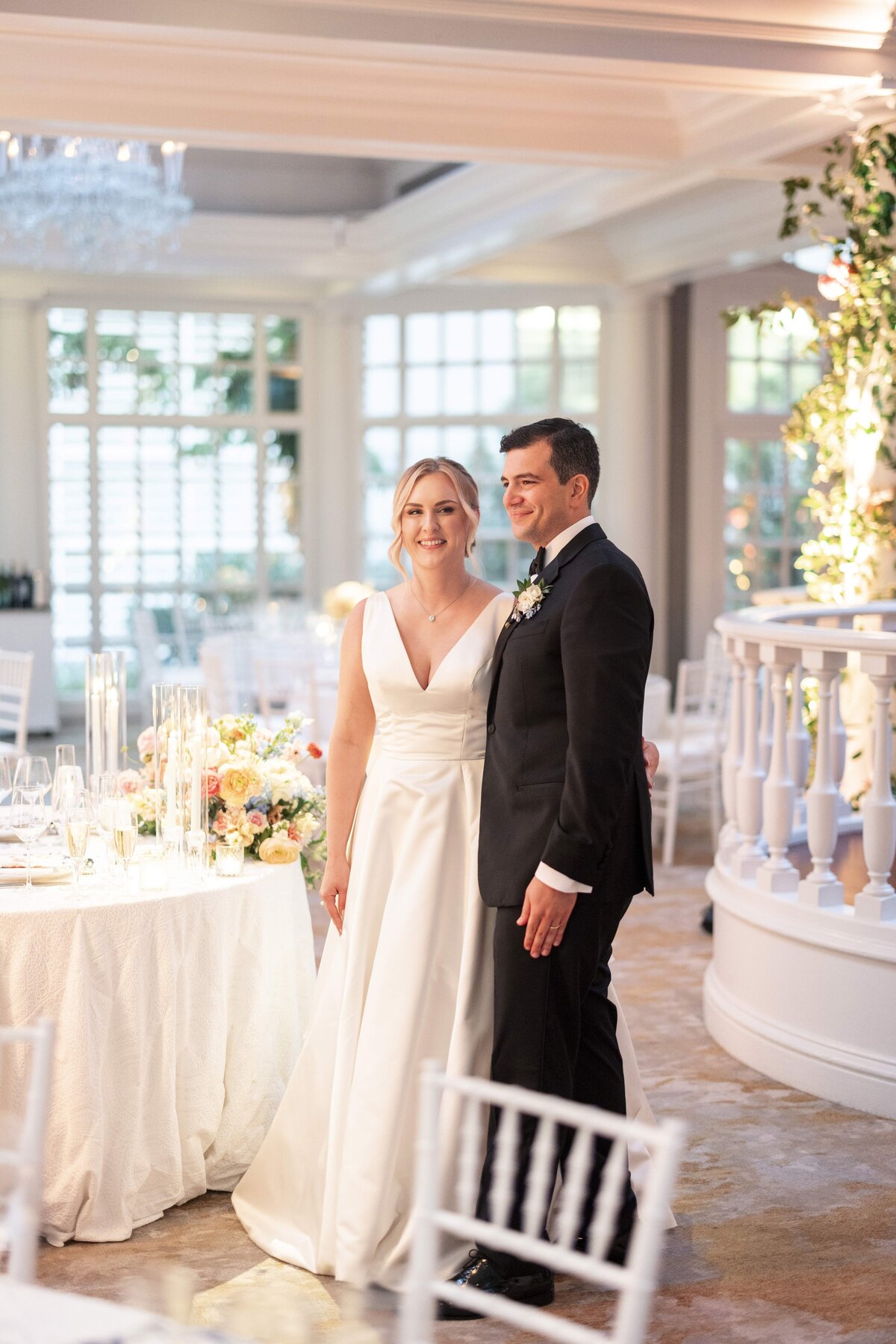 A couple dressed in wedding attire stands together in an elegant venue with white decor. The bride wears a white dress, and the groom is in a black suit. They are smiling, standing beside a table with candles and flowers.