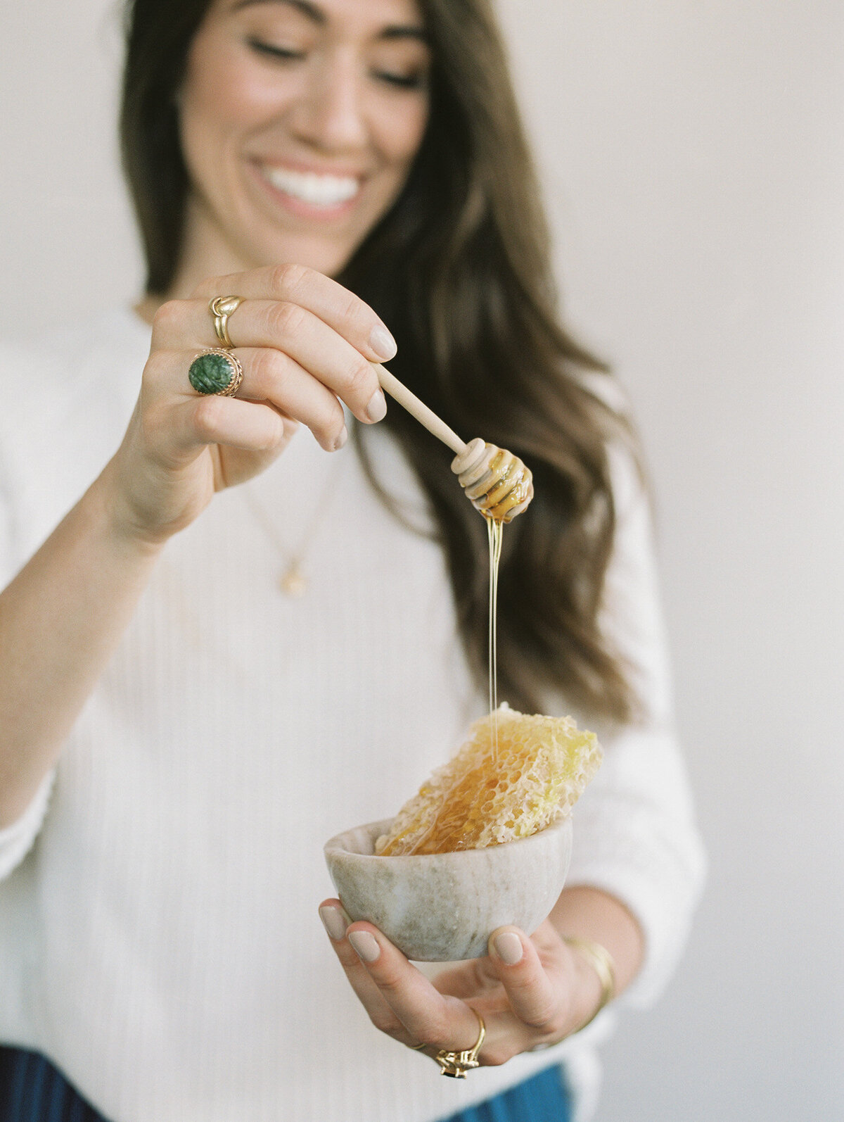 smiling woman holds honey dipper covered in honey over honeycomb in bowl