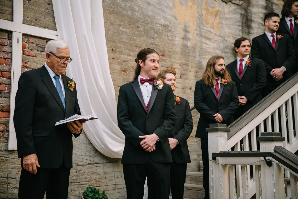 A group of groomsmen in black suits with red ties and boutonnieres stand on a staircase at a wedding ceremony. An officiant holding a book stands nearby against a rustic brick wall with draped white fabric.
