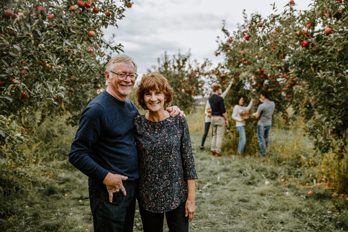 Extended family multigenerational photoshoot at apple orchard in London, Ontario. The parents, in their 50's, are standing together in the foreground smiling at the camera. Their adult children can be seen picking apples behind them in the distance.