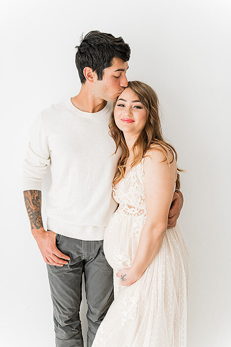 Partner kisses the expecting mom’s forehead during a maternity session in soft, neutral tones inside a white studio.