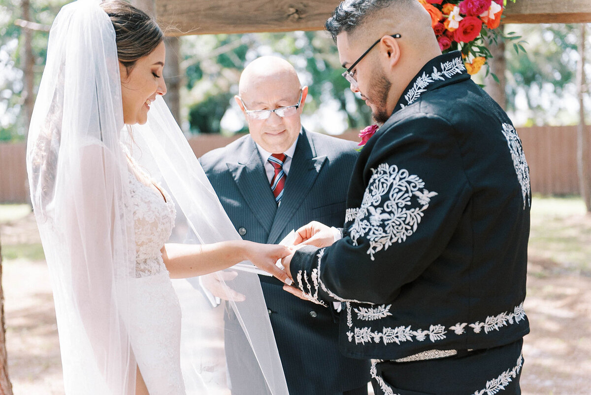 Mexican wedding with bride in a mermaid lace dress and a long veil smiling as her groom slips a wedding ring on her finger during their Tampa Florida wedding ceremony
