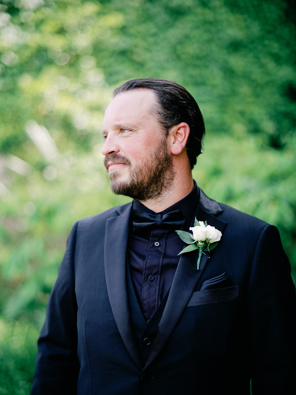 Portrait of groom wearing all black and one white flower on his jacket as he looks sideways with lush green in the background.