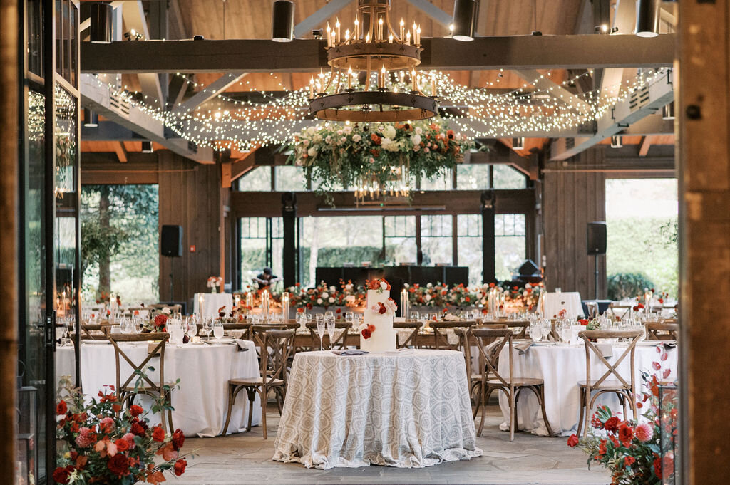Luxury wedding reception with tiered cake, red floral arrangements and large hanging chandelier installment.