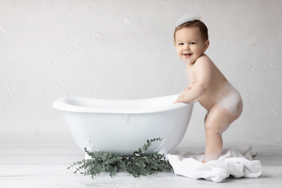 Dallas milestone photographer captures a joyful baby standing next to a white tub, covered in bubbles with a sudsy crown on their head, radiating playful charm.