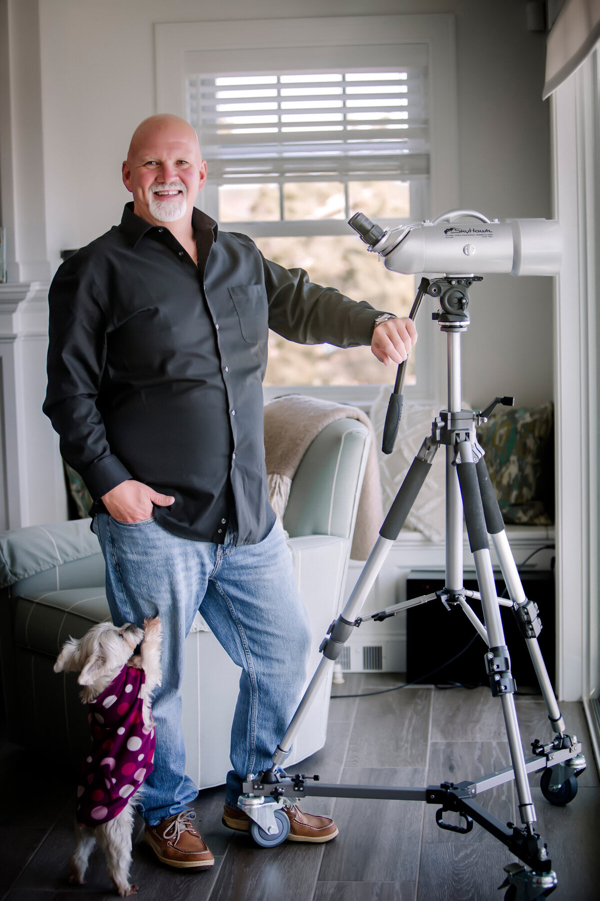 A male dating portrait client standing in his living room next to a telescope, and his small dog jumping on him