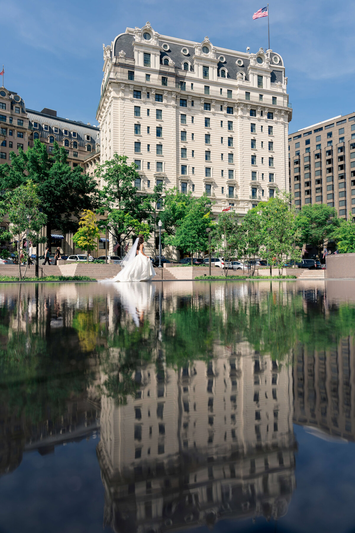 A grand historical building with ornate architecture reflected in a calm body of water. A couple, with a bride in a white dress, walks along a path in front of the building, surrounded by green trees, under a clear blue sky.