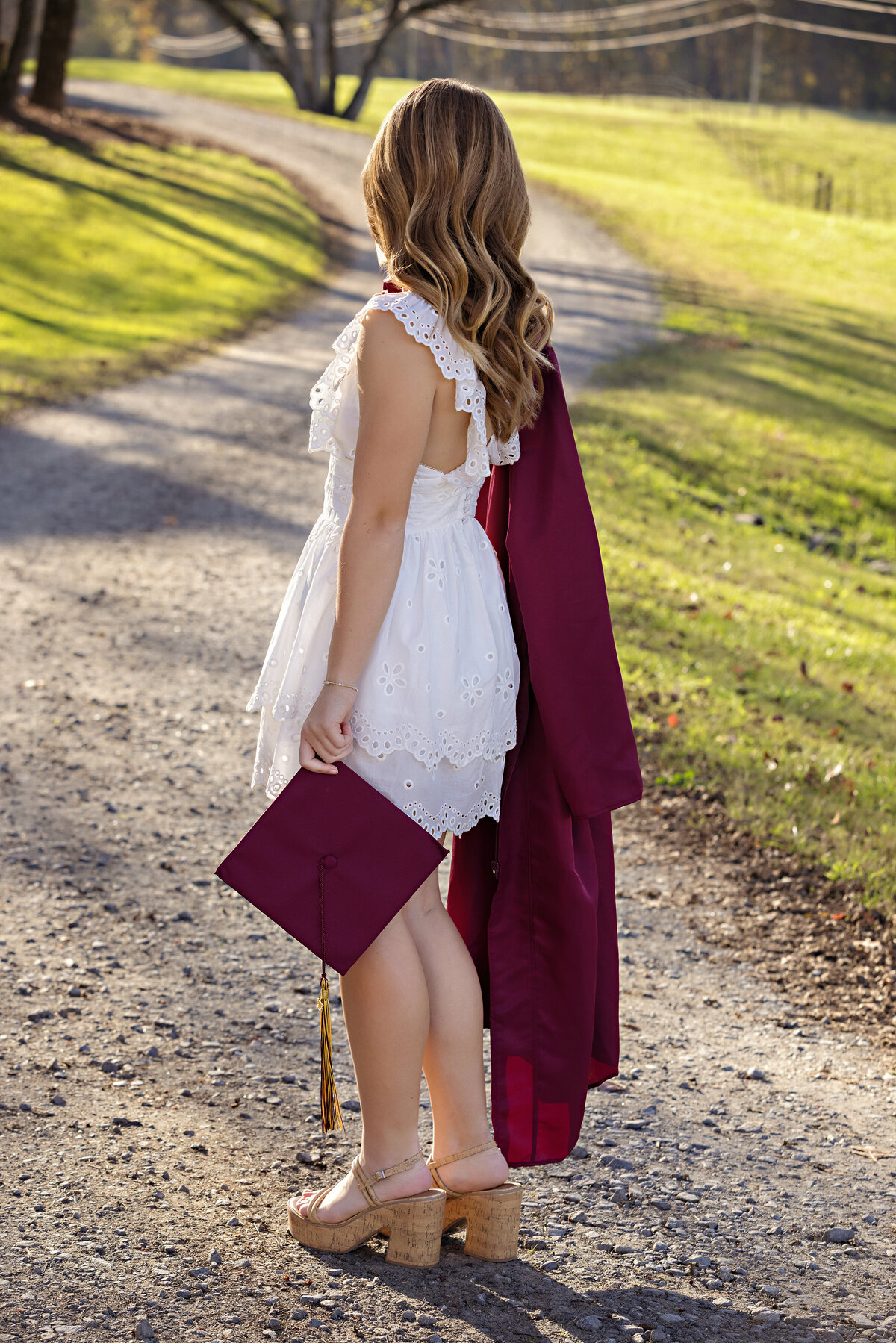 High school senior in cap and gown near gravel road with golden light filtering through trees
