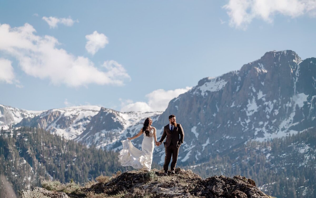 A bride and groom enjoy a sunny, windy day on front of snow capped mountains.