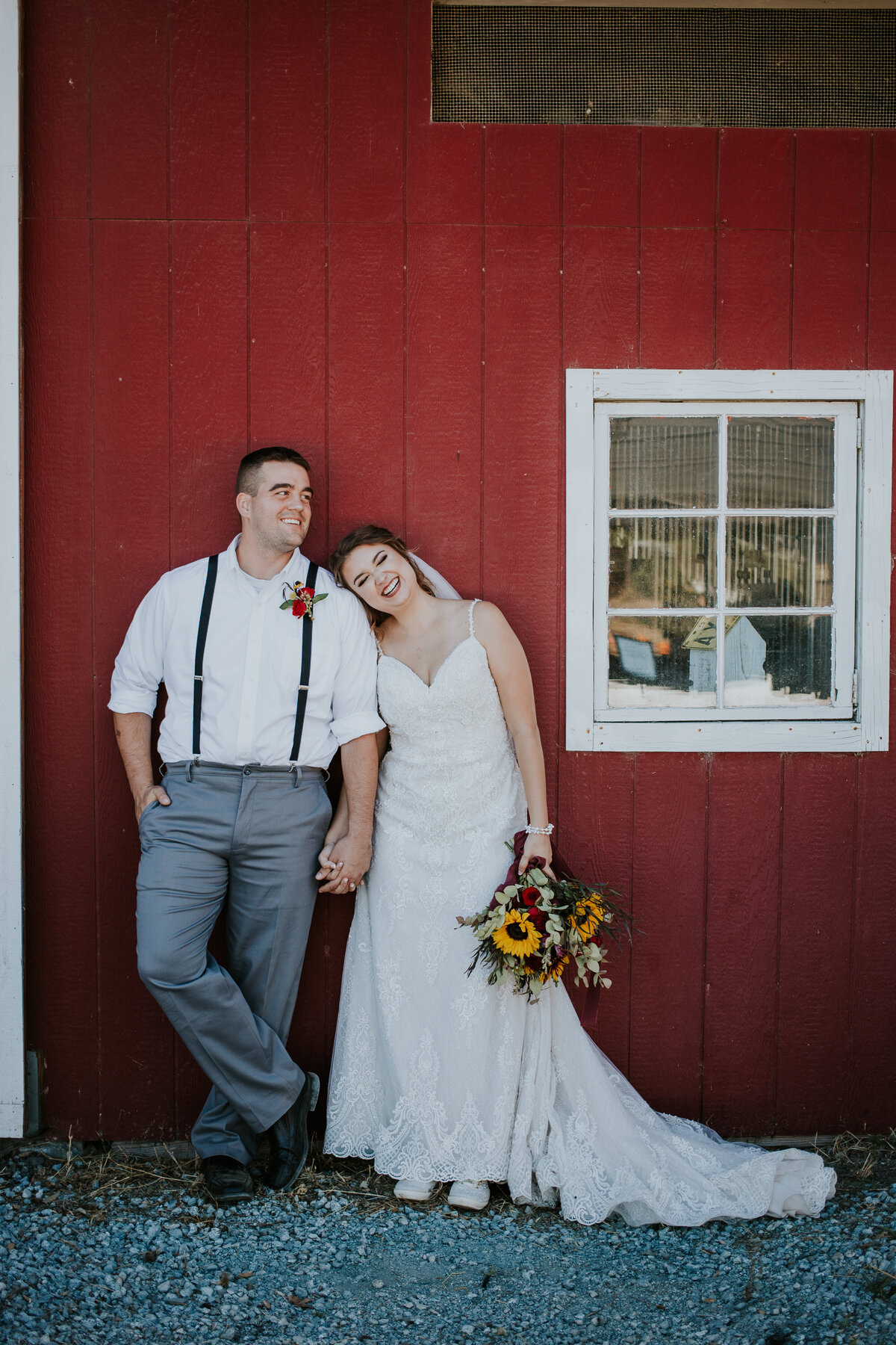 Standing in front of red barn, bride leans on grooms shoulder as they hold hands and smile.