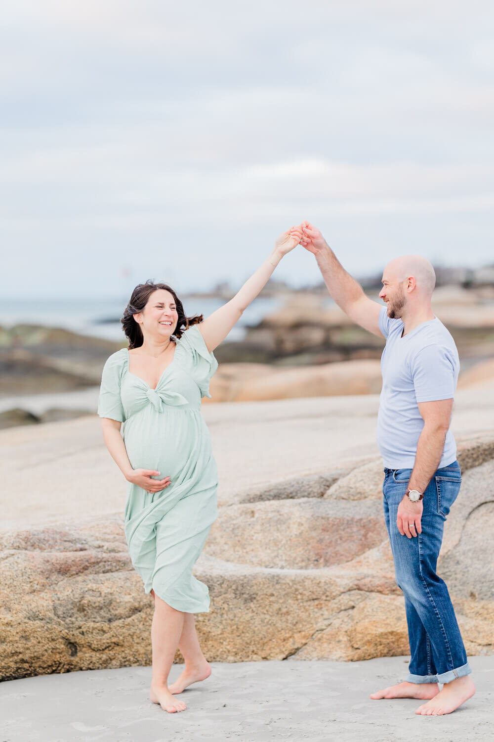 Man twirls his smiling, pregnant wife while dancing on a beach