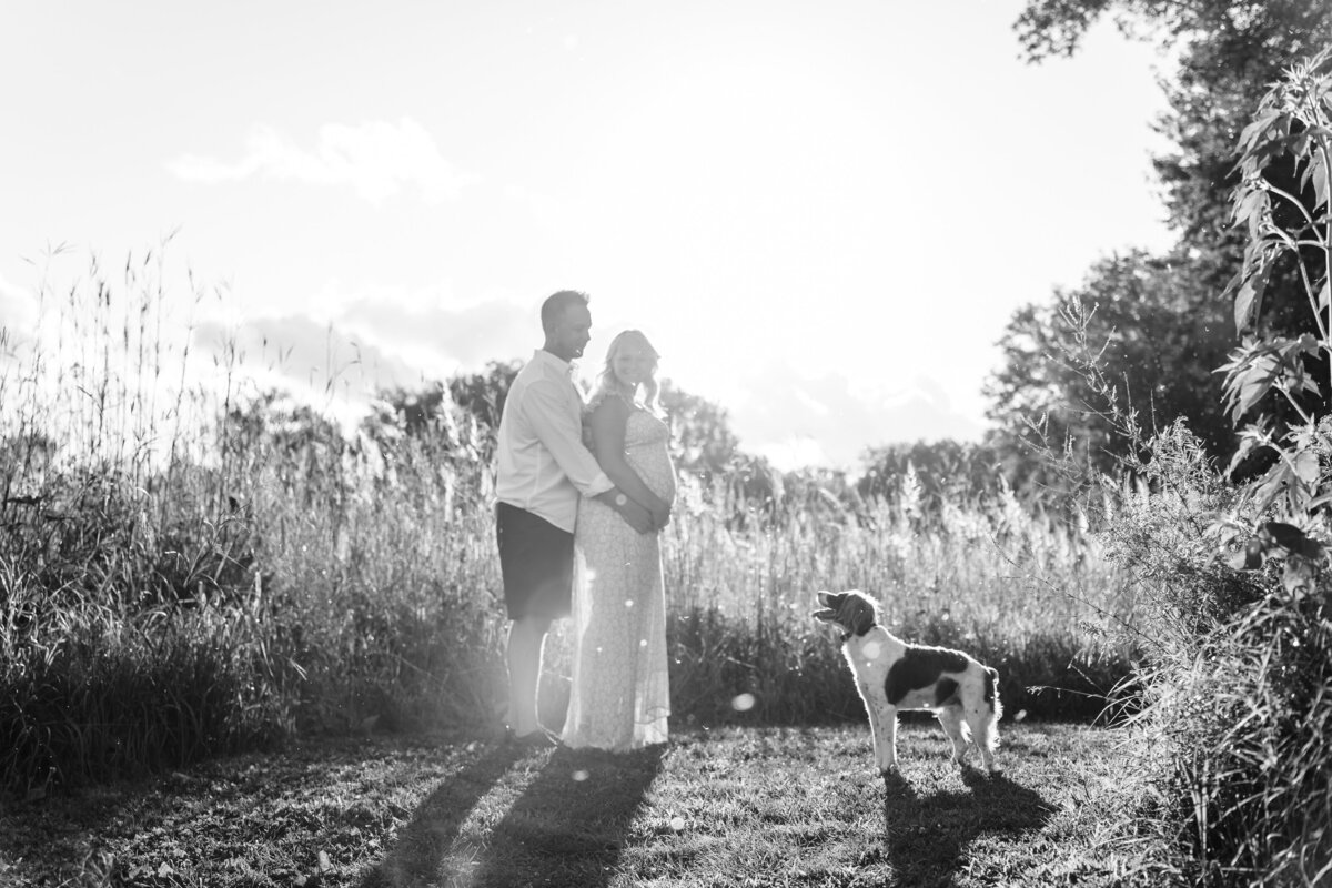 A black and white image of the couple standing in a grassy field with their dog. The man is holding the woman from behind, both looking out into the distance.